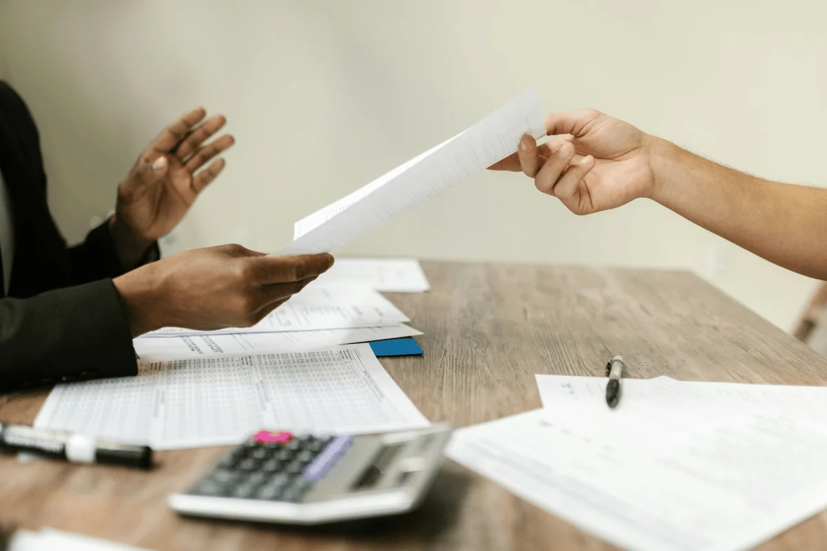 Two people exchanging a document across a table with papers and a calculator.