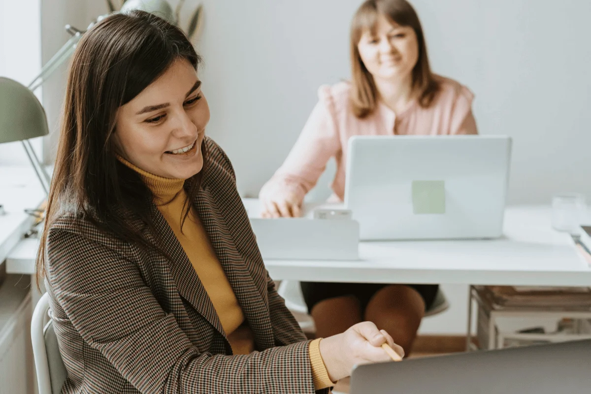 Two women collaborating in an office setting, smiling while working on laptops.