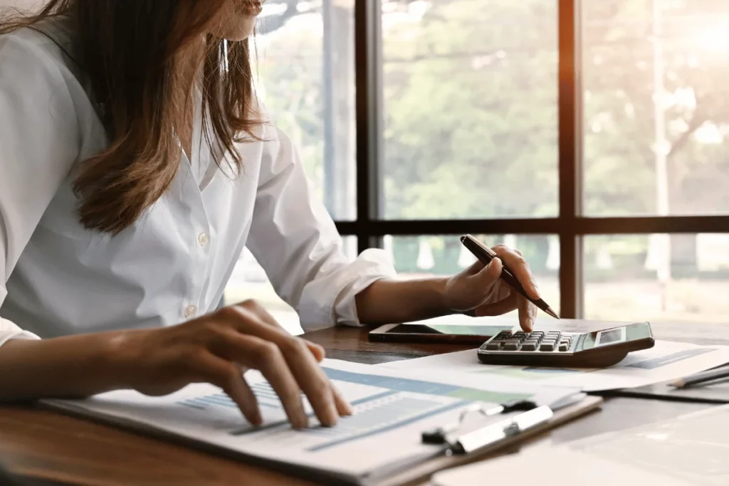 Woman analyzing graphs with calculator at desk near window.