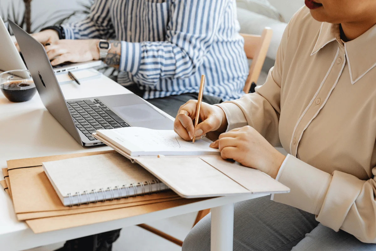 Person writing on paper next to a laptop with others working in the background.