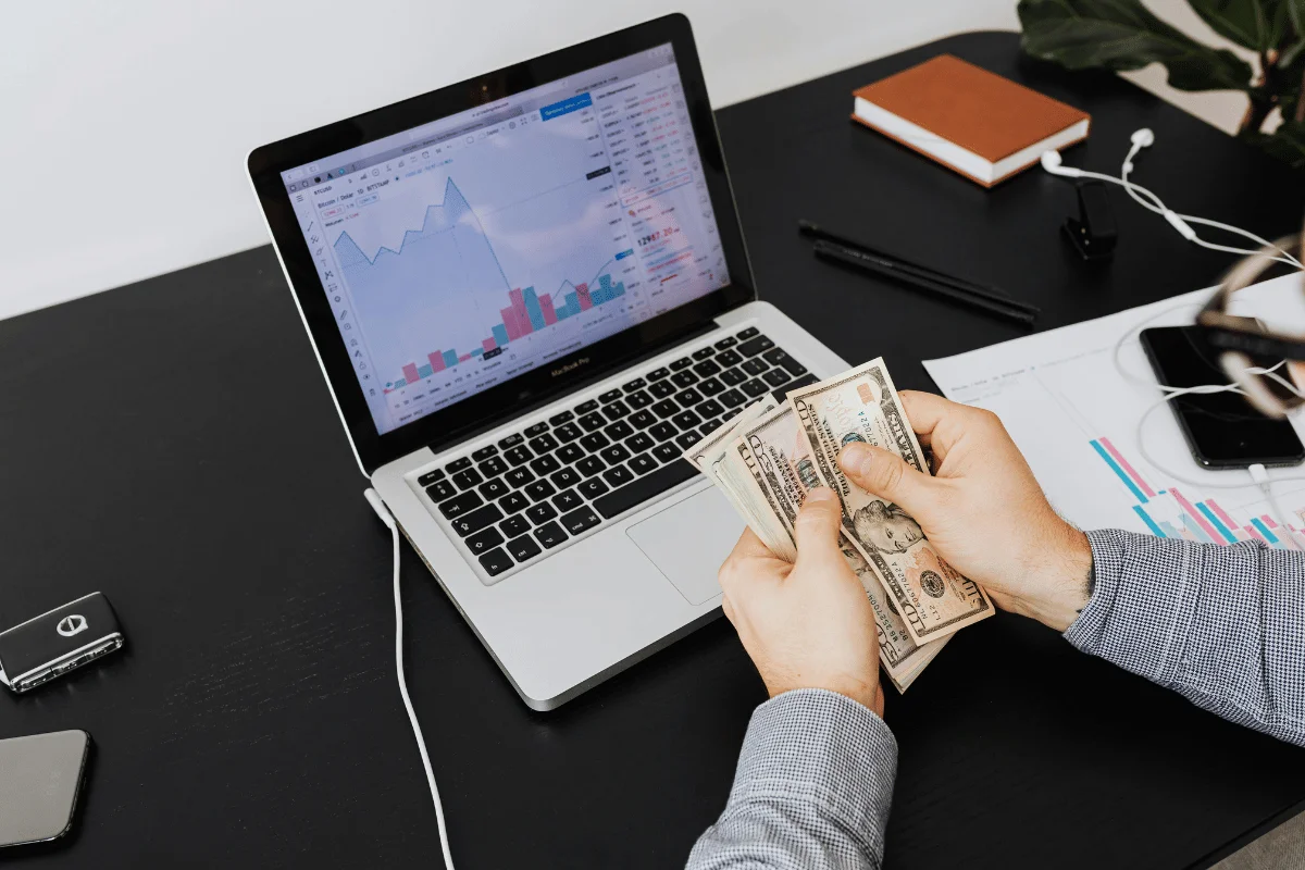 Hands counting dollar bills in front of a laptop displaying financial charts on the screen.