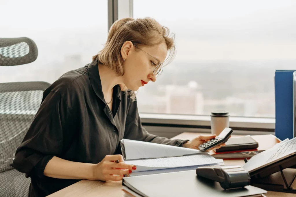 A person in a black shirt is working at a desk with papers, a calculator, and a coffee cup, overlooking a city skyline.