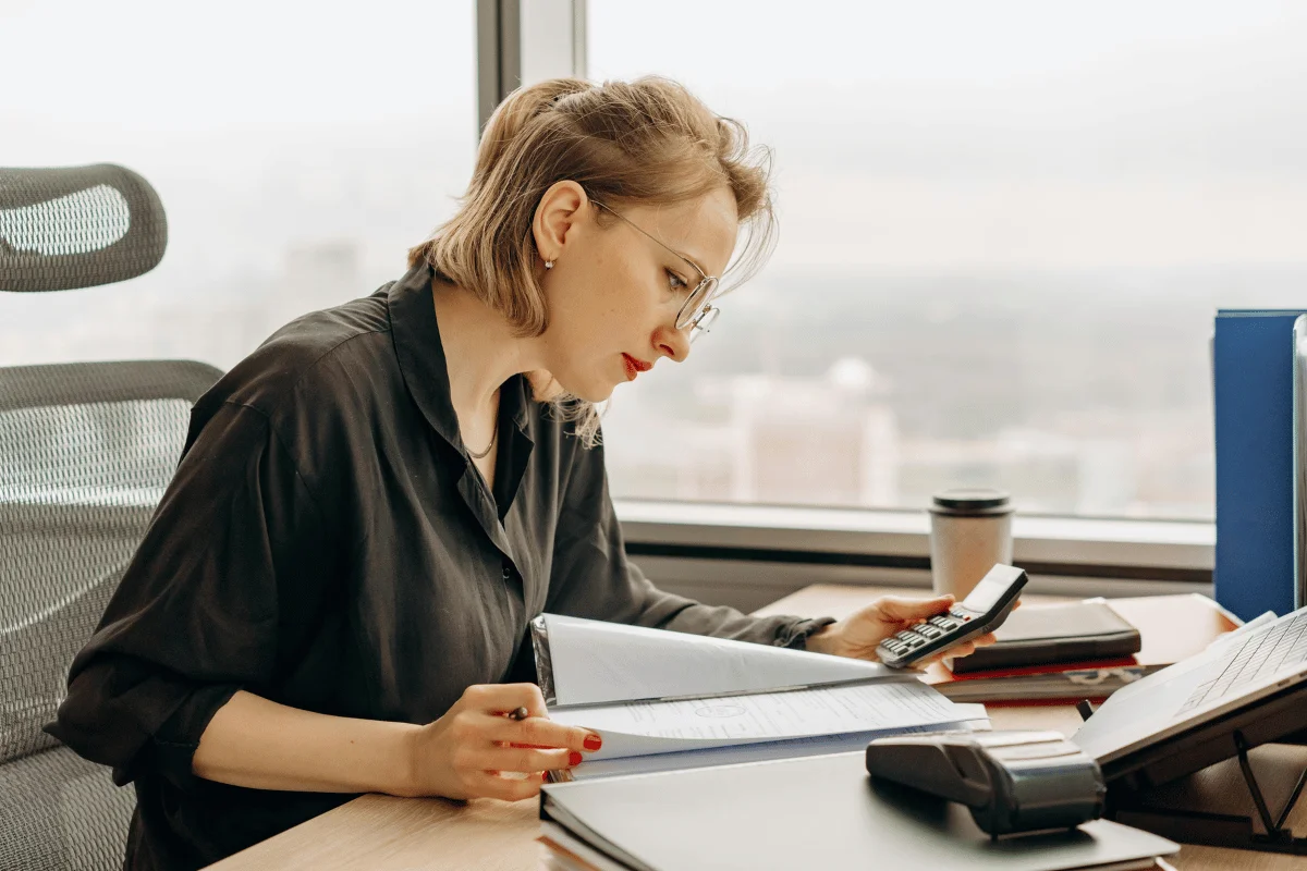 A person in a black shirt is working at a desk with papers, a calculator, and a coffee cup, overlooking a city skyline.