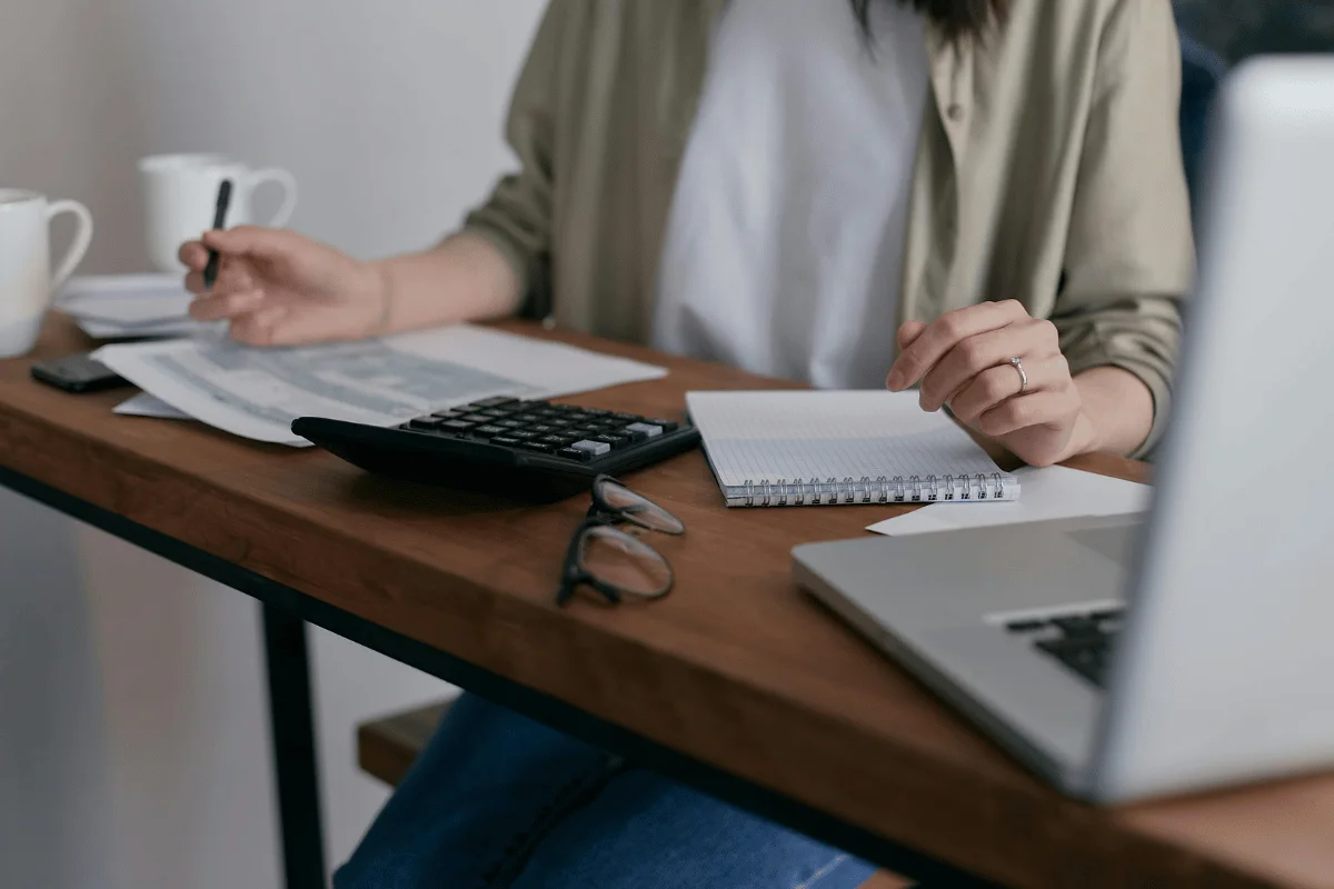 A person reviews documents and uses a calculator at a wooden desk, with a laptop and coffee mugs in a cozy workspace.
