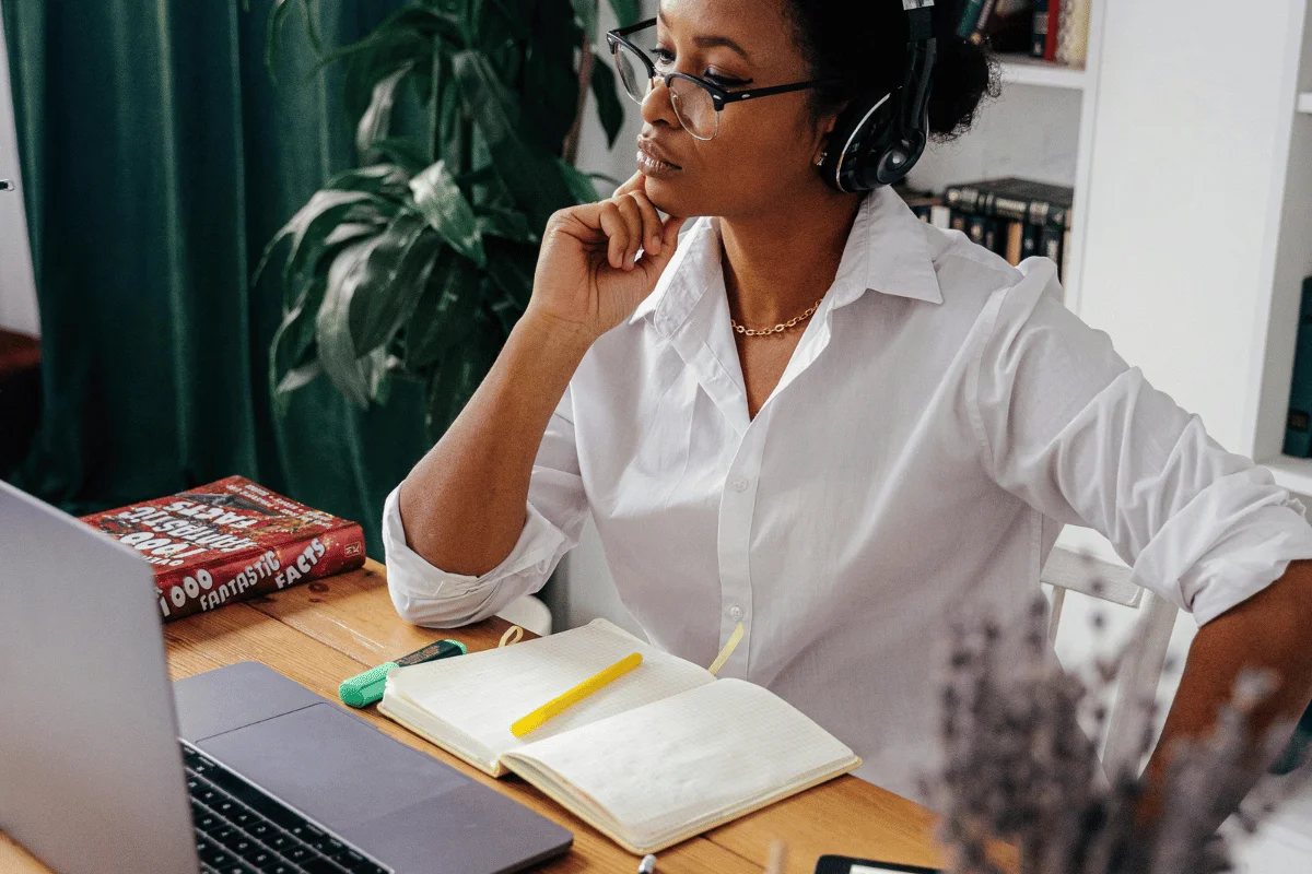 A person in a white shirt with headphones sits at a wooden desk with a laptop and an open notebook, deep in thought.