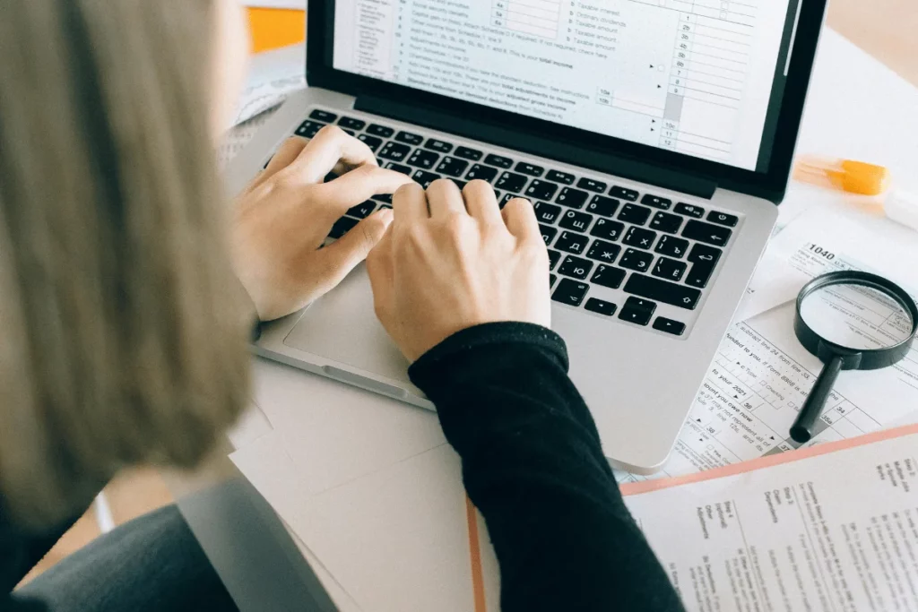 A person typing on a laptop with financial documents and a magnifying glass on a desk, focused on data entry.
