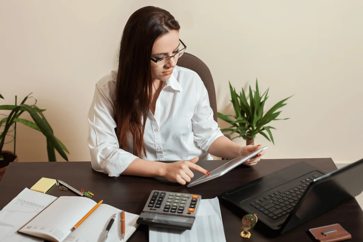 Businesswoman working at a desk using a tablet, surrounded by documents, a calculator, and a laptop.
