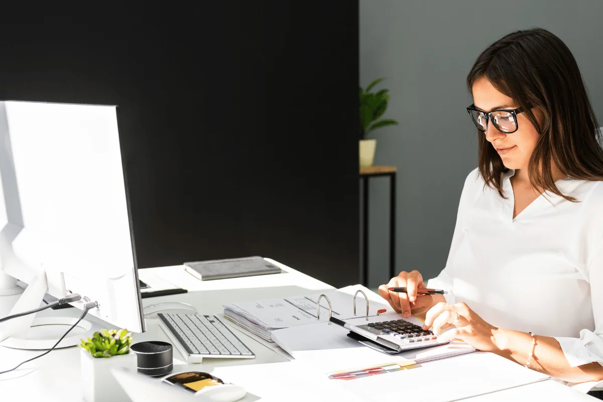 Businesswoman analyzing financial data with a calculator and files in a well-lit office.