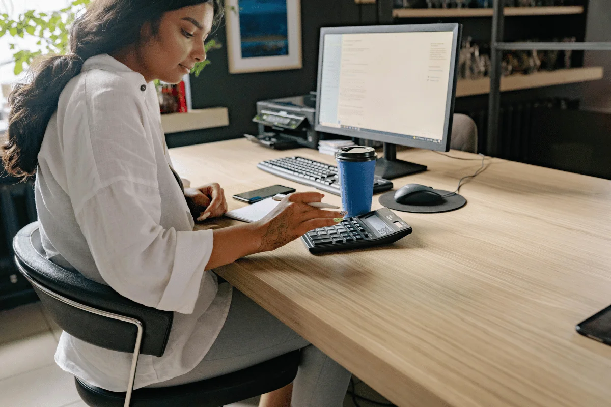 A woman working at a desk with a computer, calculator, notebook, and coffee cup.