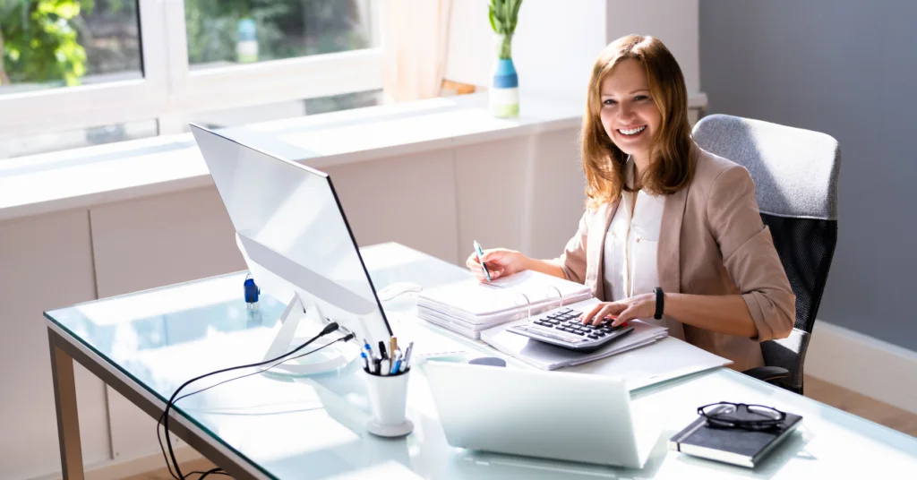 Woman working at a desk with a computer, laptop, calculator, and notepad.