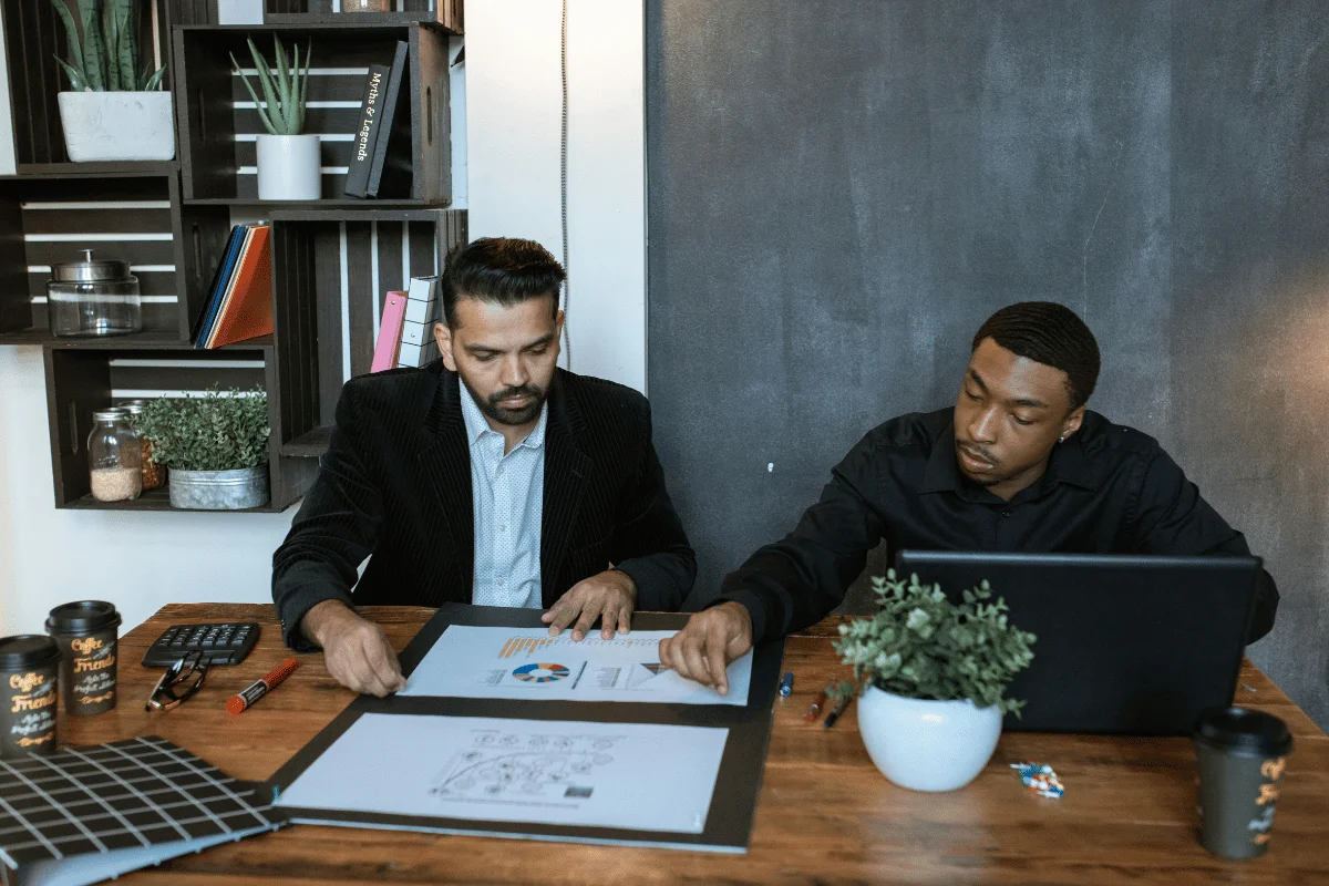 Two people at a desk with documents and a laptop, in a room with a bookshelf.