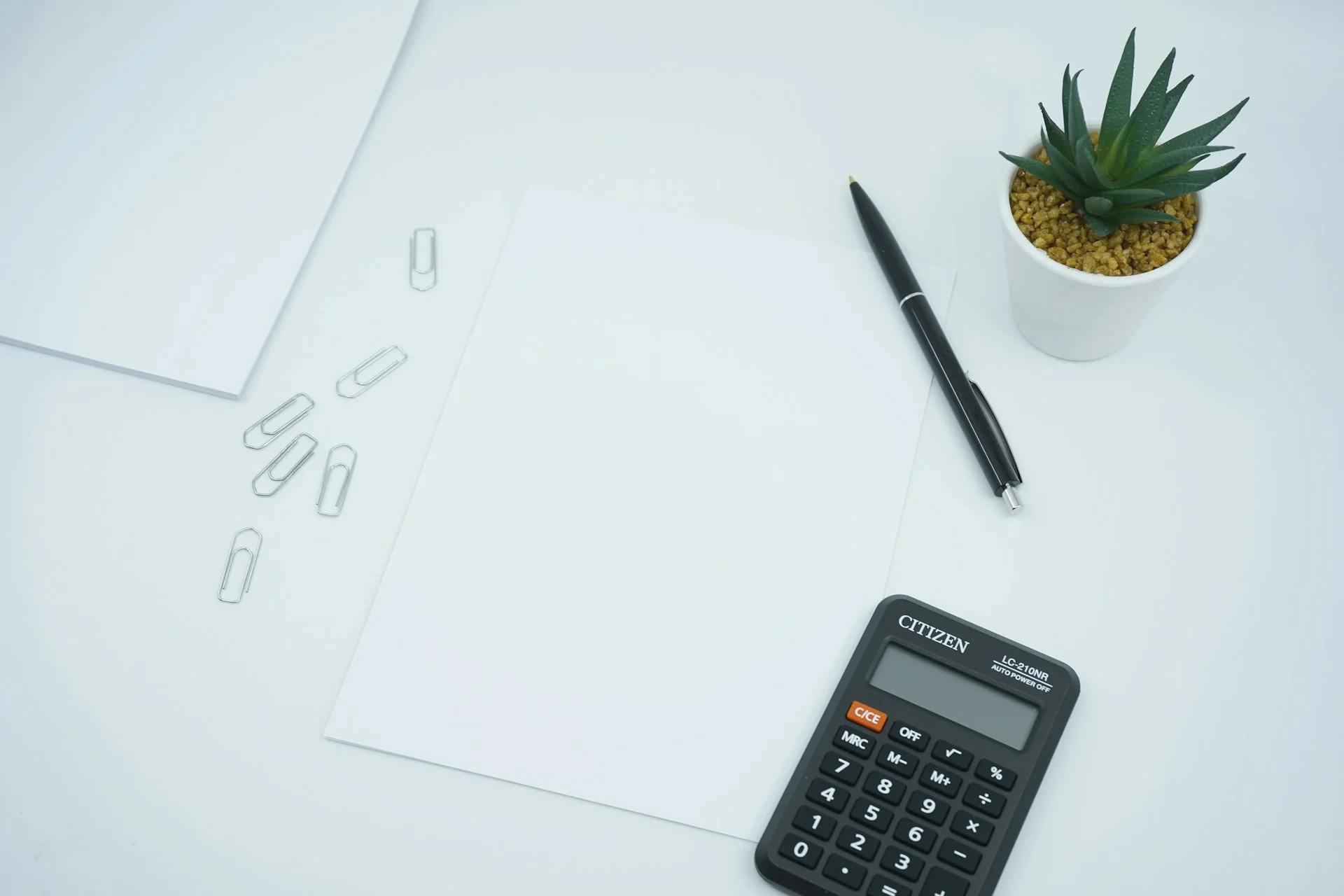Desk setup with a calculator, blank papers, pen, and a small potted plant.