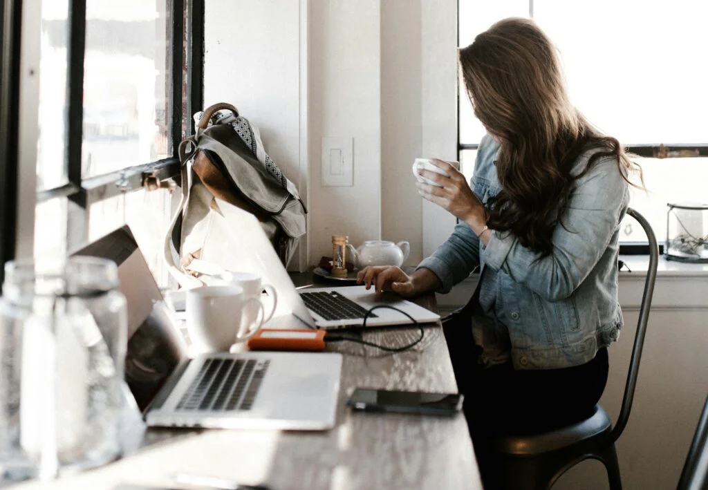 A woman holding a coffee cup and scrolling on a laptop.