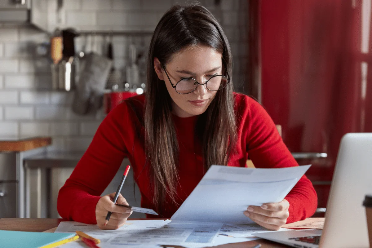 A woman in a red sweater looking at documents while holding a pen and sitting at a table with a laptop nearby.