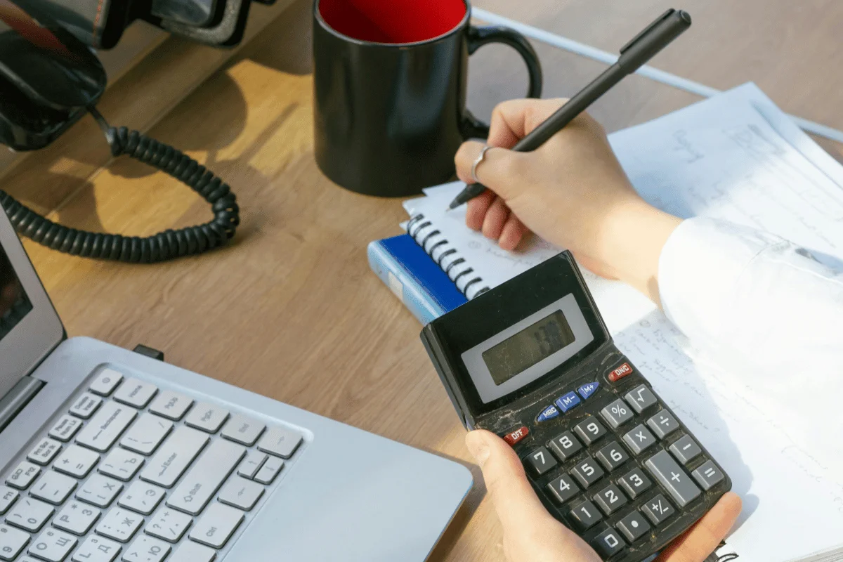 A person holds a calculator in one hand while writing in a notebook with a pen on a desk.