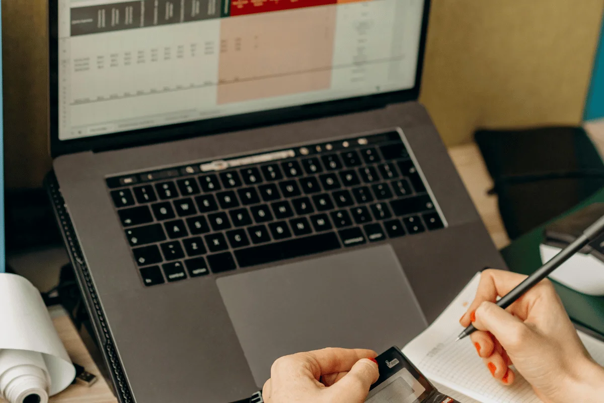 A person using a calculator while writing in a notebook with a laptop displaying financial data in the background.