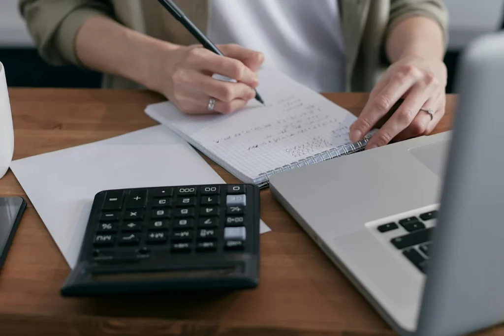 Person writing financial notes with calculator and laptop on desk.