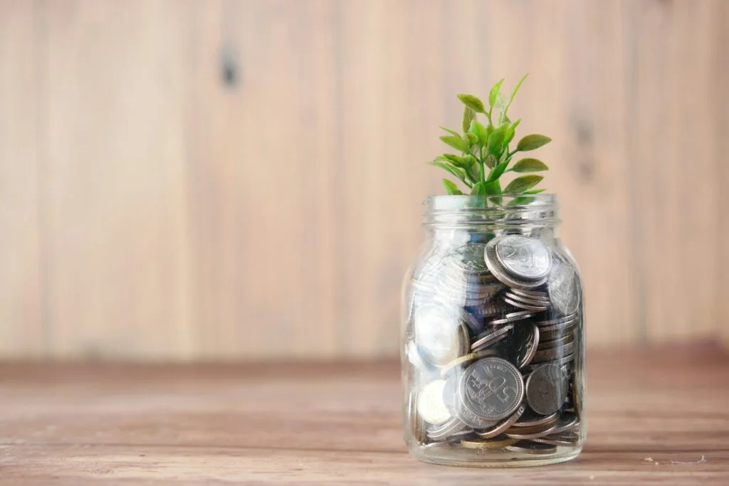 A glass jar filled with coins, topped with a small green plant, sits on a wooden surface. The image conveys growth and saving concepts.