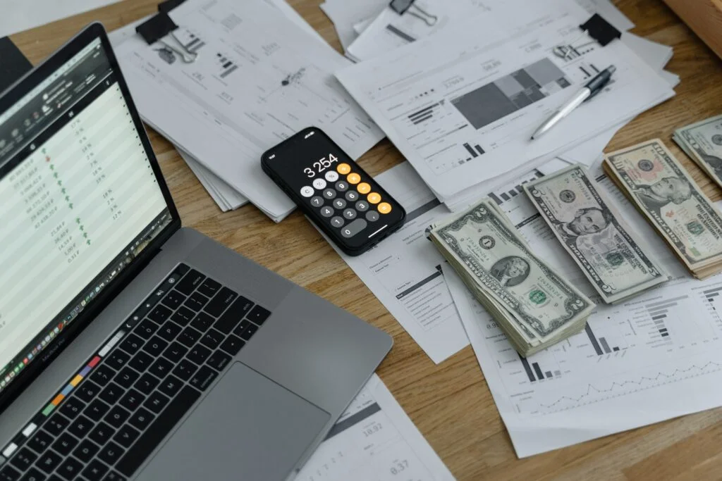 A laptop displays financial data next to a calculator showing $3,254 and stacks of cash on analysis papers, depicting budgeting or investing.