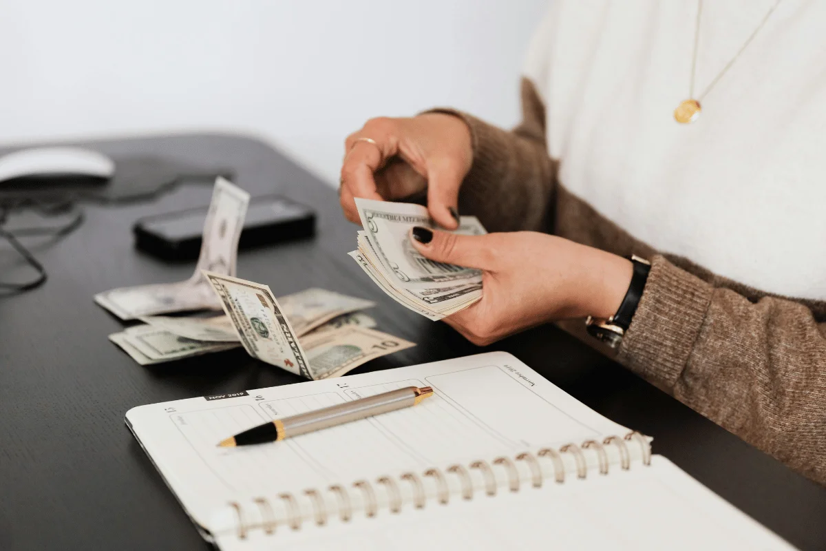 Person counting US dollar bills at a desk with a notebook and pen.