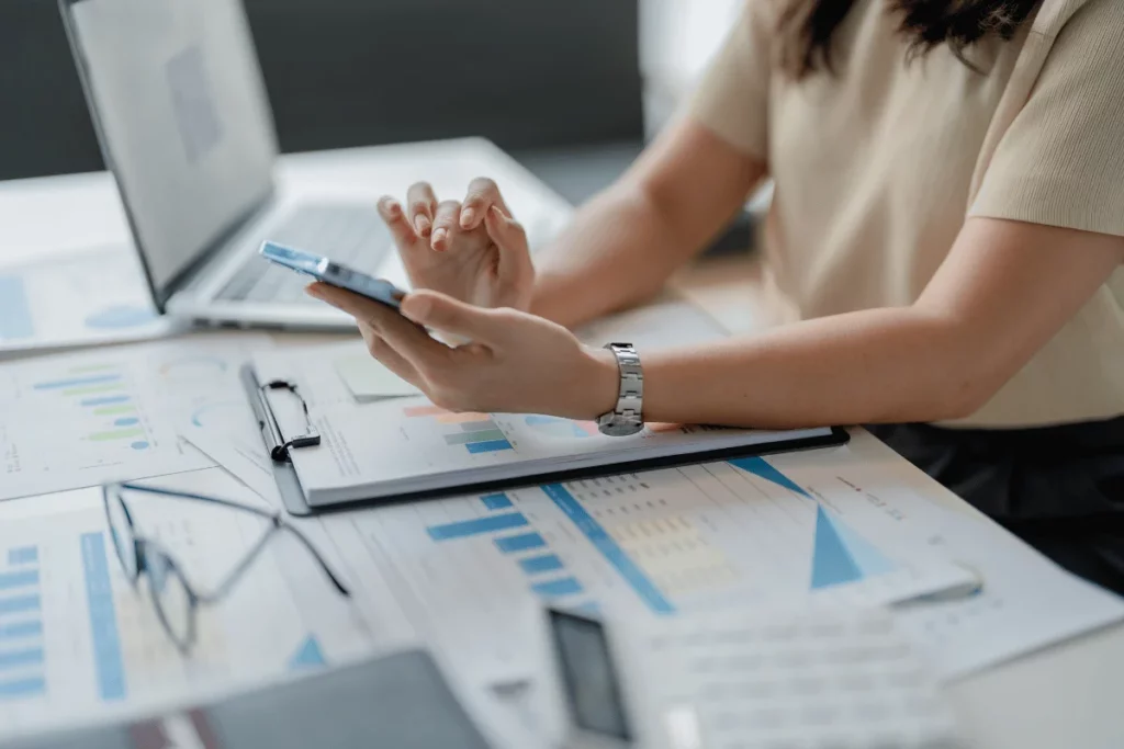 Businesswoman analyzing financial charts and using a smartphone at her desk.