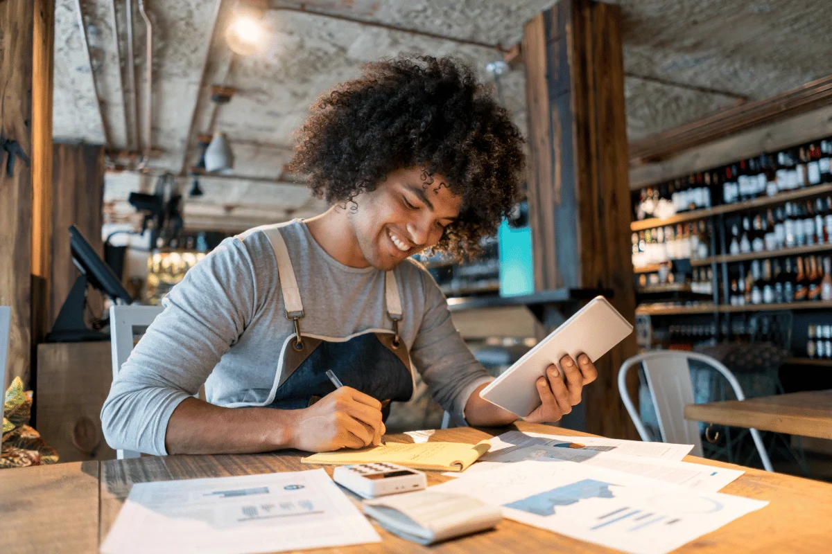 Entrepreneur working on financial plans using a tablet and notepad in a coffee shop.
