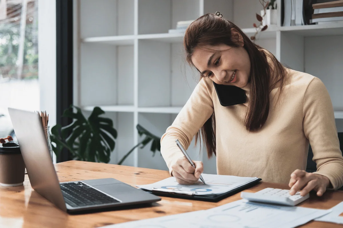 Woman multitasking at desk, using calculator and talking on phone.