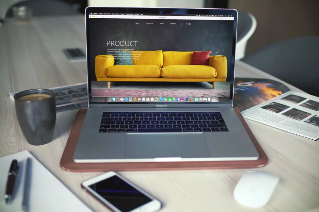 A laptop on a desk surrounded by a cup of coffee, pen and paper, a smartphone, a newspaper, and a magazine.