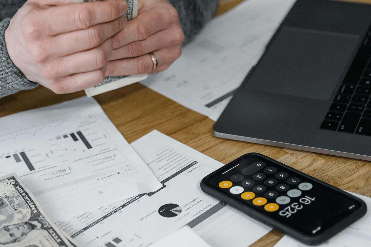 Person counting cash with financial documents and a calculator app open on a phone.