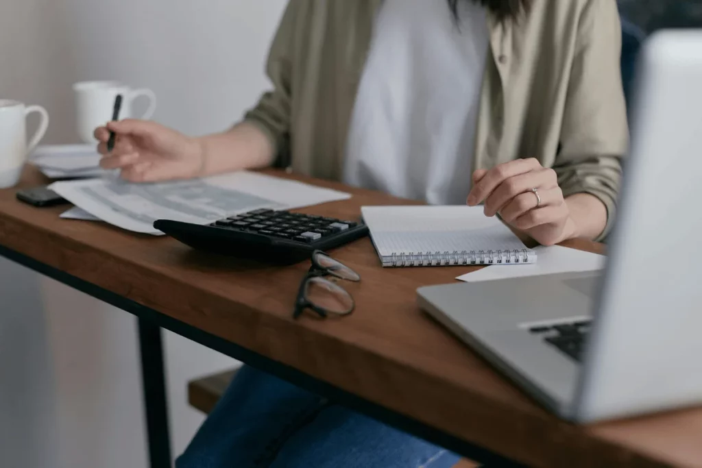 Person working on financial documents with a calculator and notebook at a wooden desk.