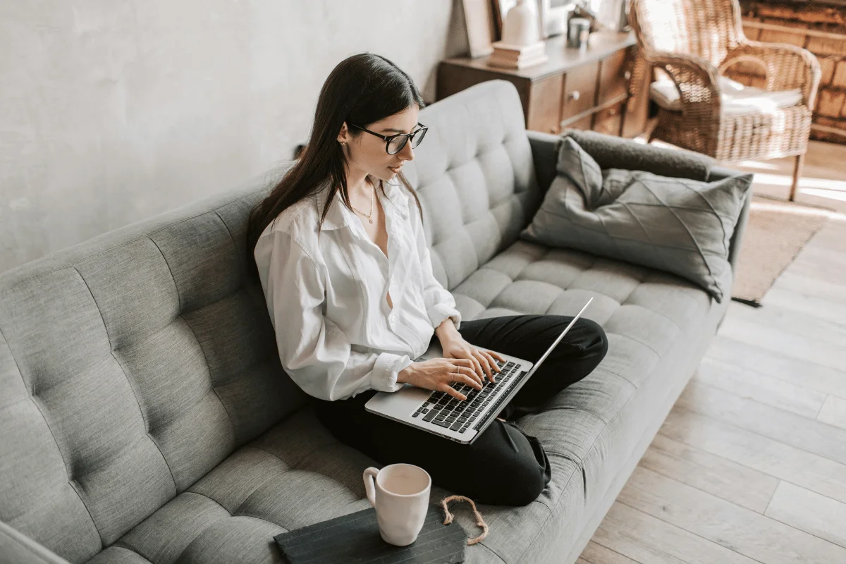 Woman working on a laptop while sitting on a grey couch in a cozy living room.