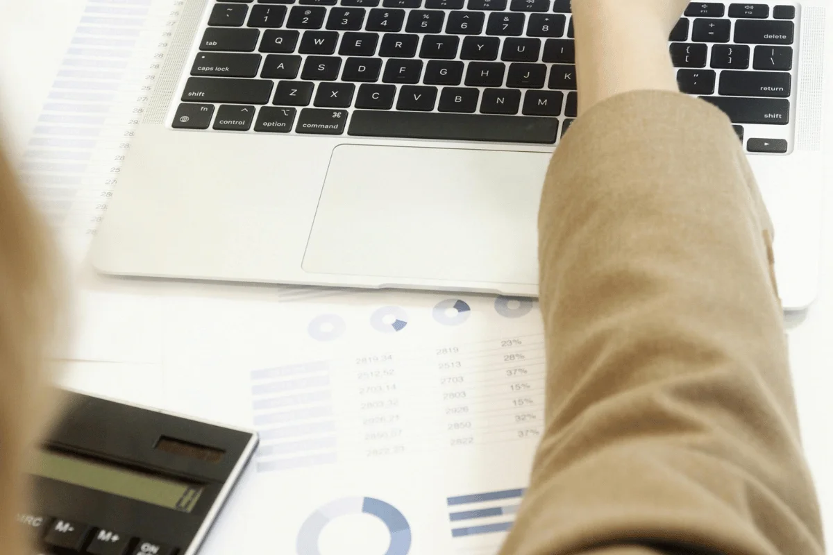 Individual typing on a laptop keyboard while reviewing business reports and data charts spread across the desk.