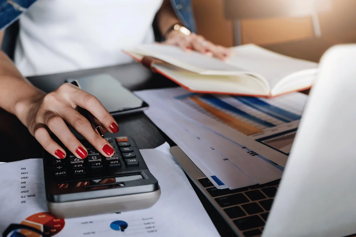 A person using a calculator while reviewing financial documents and notes, with a laptop and an open book on the table.