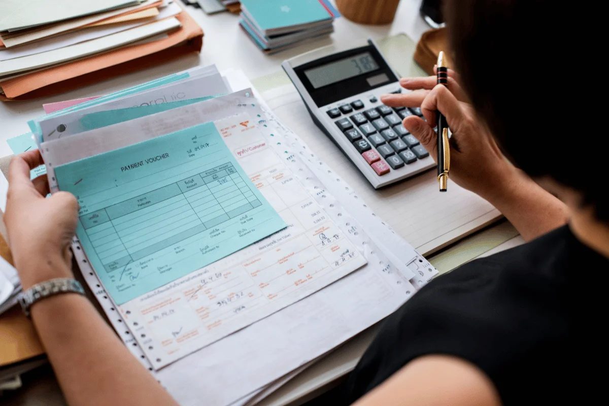 A person reviews payment vouchers and financial documents while using a calculator, preparing for accounting tasks.