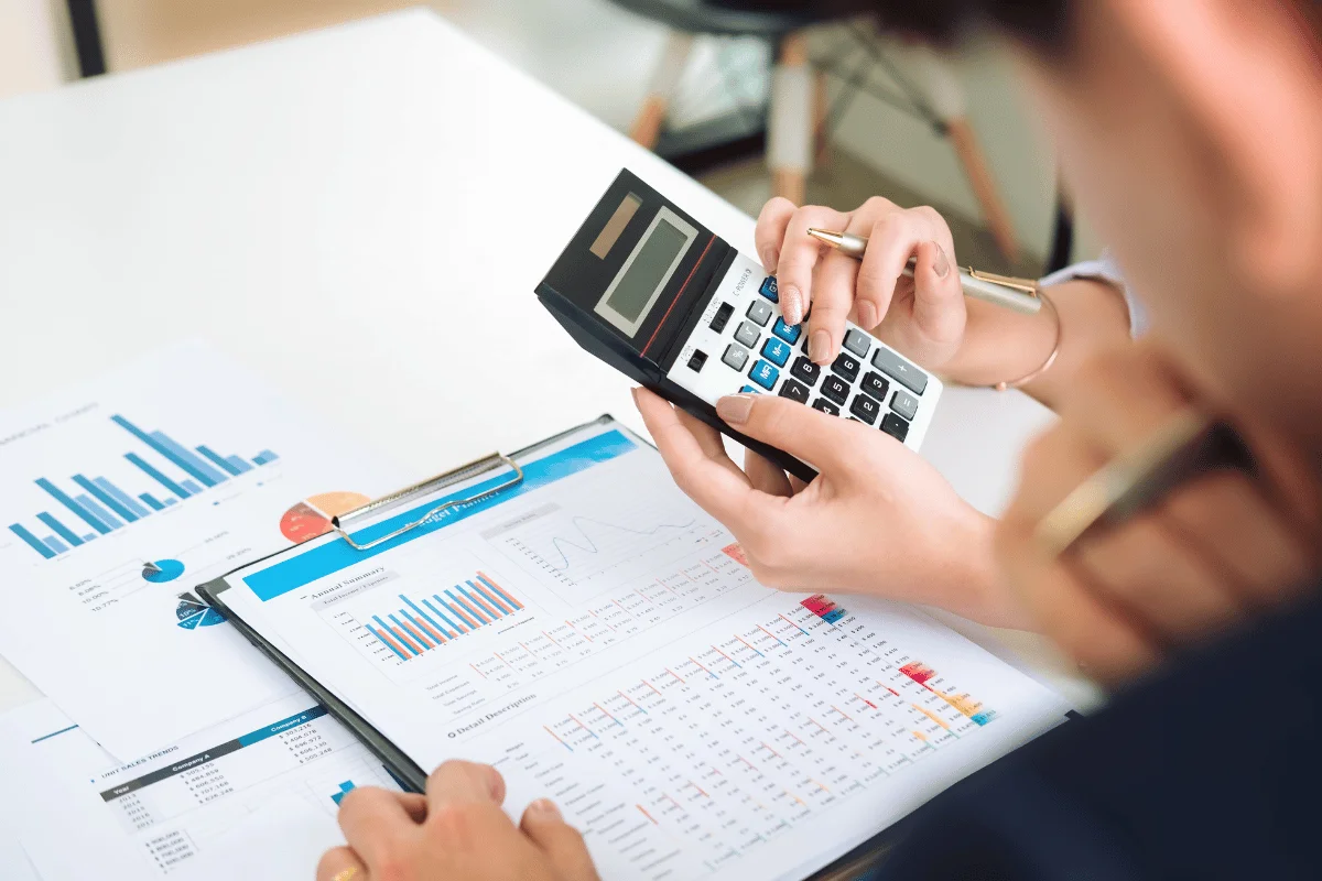 A close-up of hands using a calculator over financial documents and graphs on a desk, indicating a financial analysis or budgeting task.