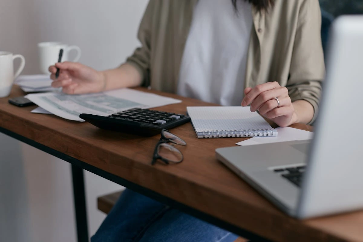 A woman is seated at a desk, working on her laptop, with a notebook placed next to her.