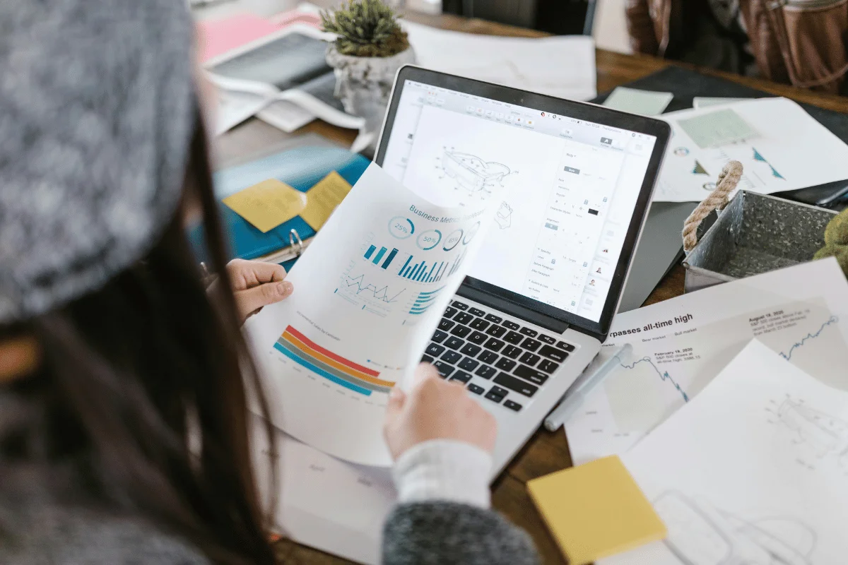 Person reviewing business reports with charts and graphs while working on a laptop, representing data analysis and business strategy.