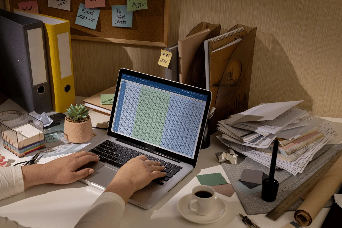 A person works on a laptop displaying a spreadsheet, surrounded by cluttered papers, a coffee cup, and colorful sticky notes.