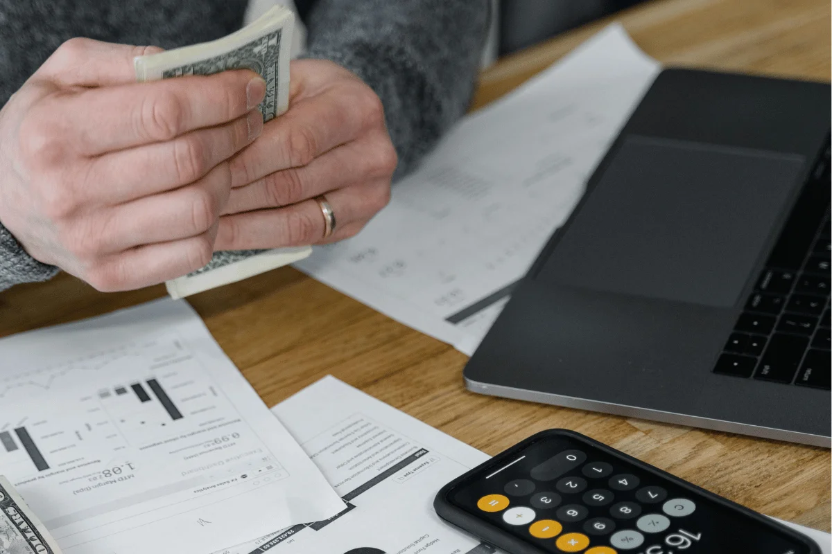 A person counting cash over financial documents and a calculator, with a laptop nearby on a wooden table.