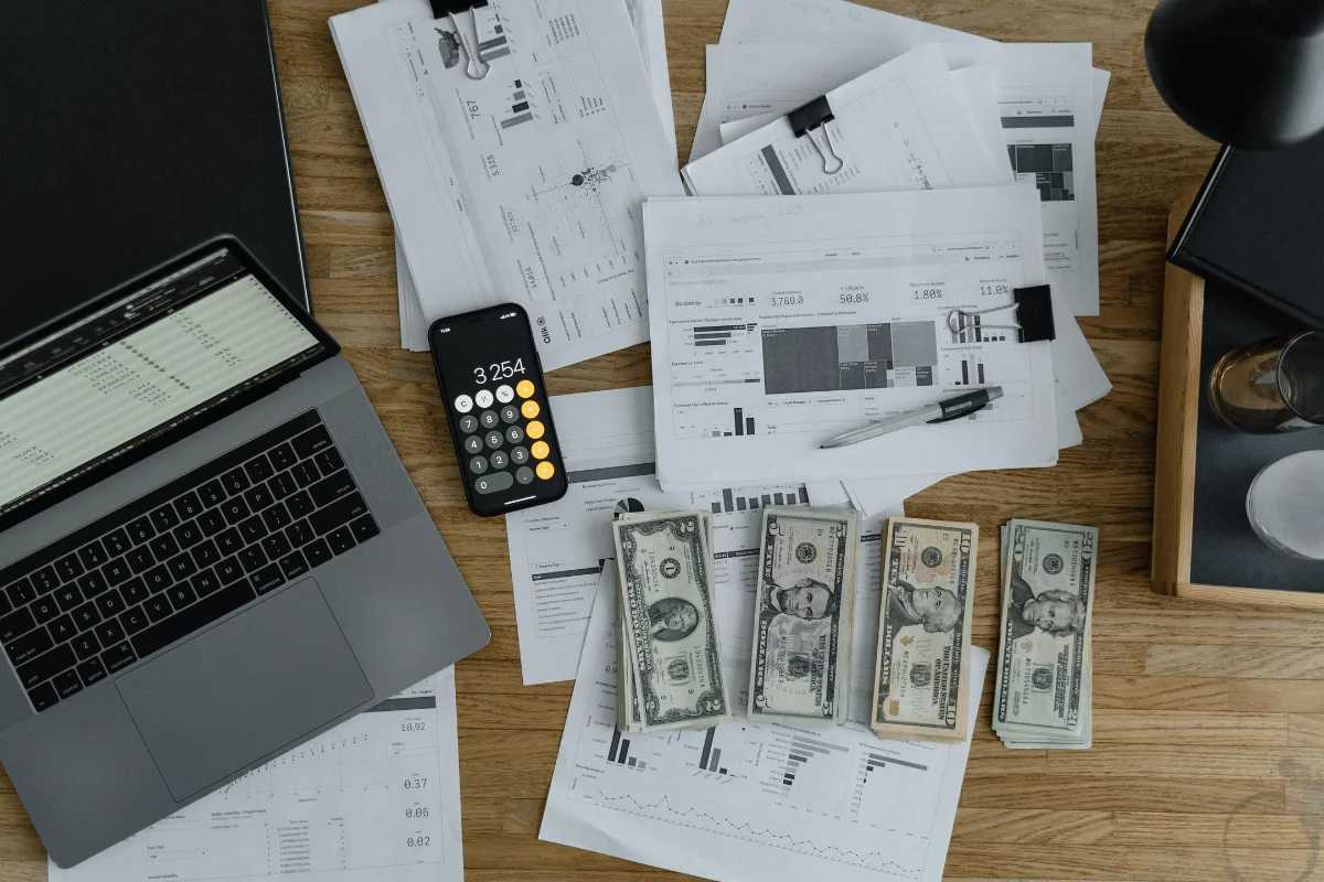 A cluttered desk with a laptop, smartphone showing a calculator app at 3,254, financial documents, and stacks of US dollar bills, conveying a busy finance-related scene.