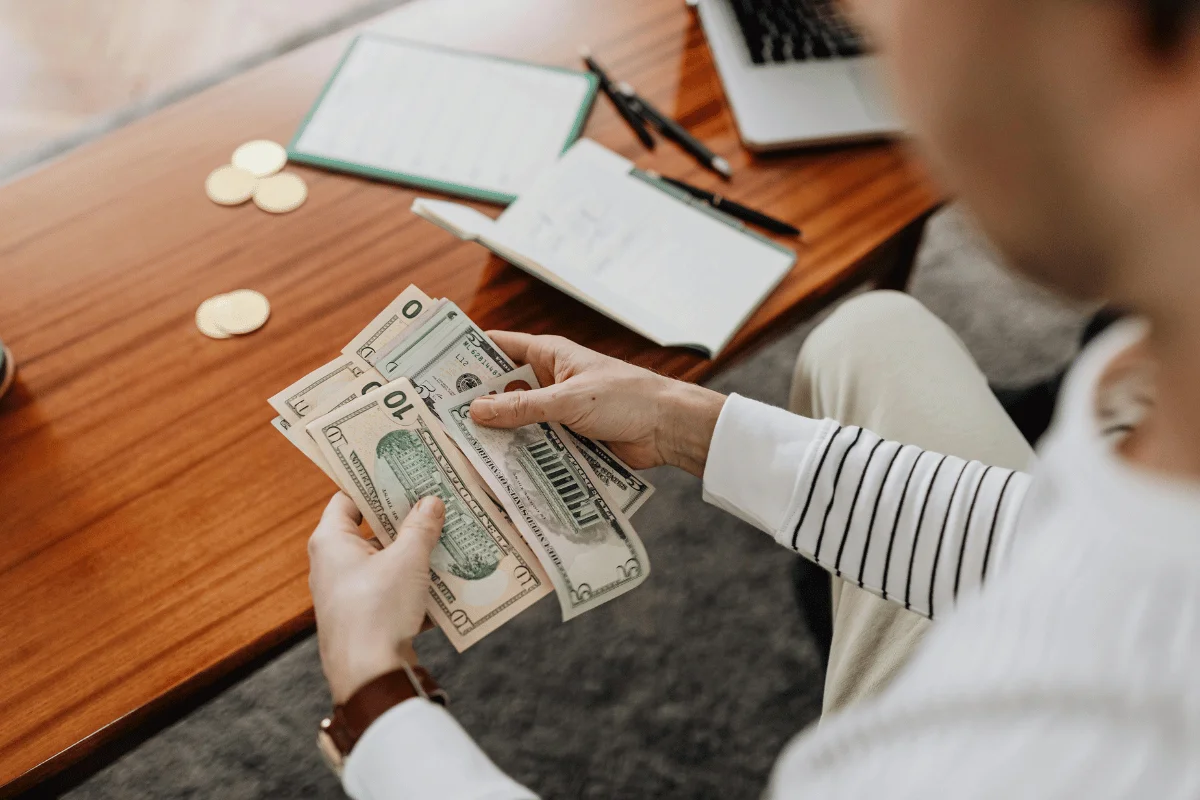 Individual budgeting and counting dollar bills with laptop nearby.