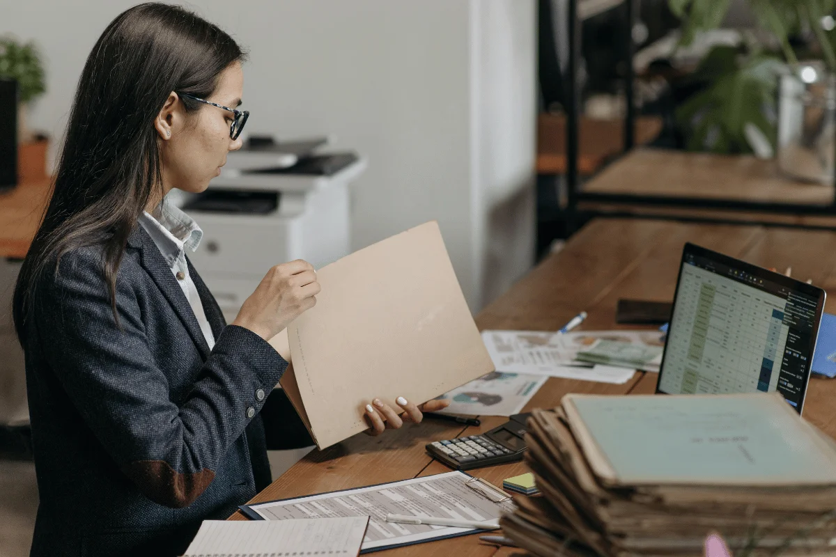 An office worker looks through a file folder while seated at a desk with a laptop and a calculator.
