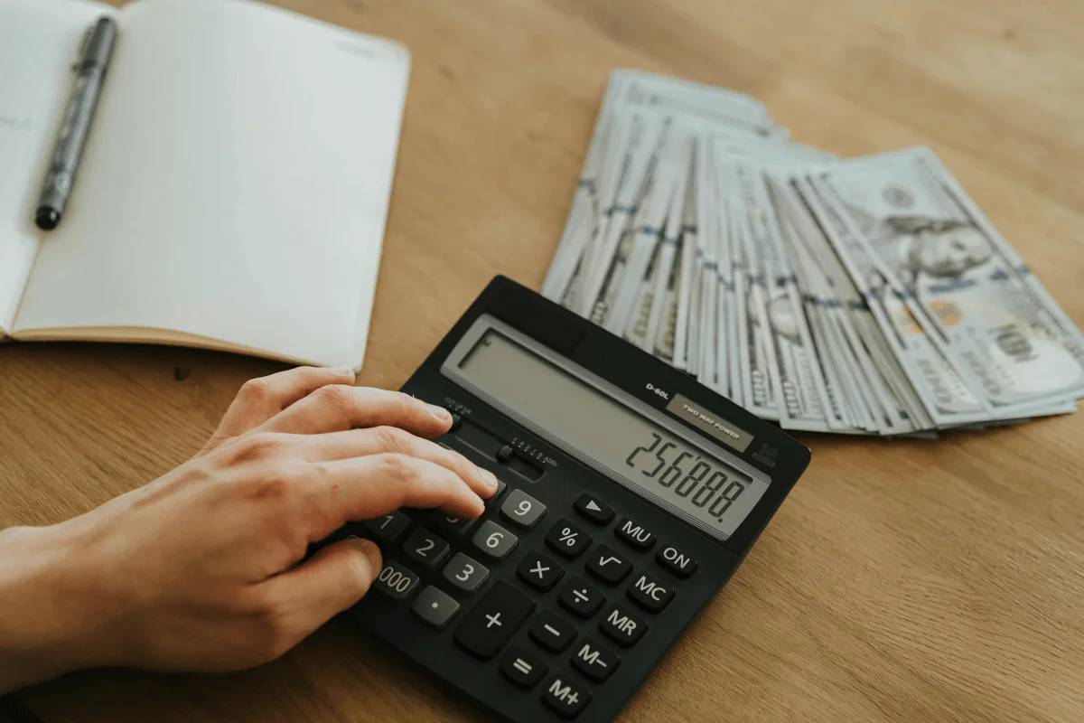 A hand using a calculator displaying 256,888, next to a notepad and a stack of neatly arranged dollar bills on a wooden table.