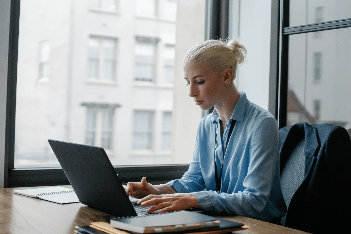 A woman in a blue shirt works intently on a laptop at a desk by a large window, with a notebook nearby.