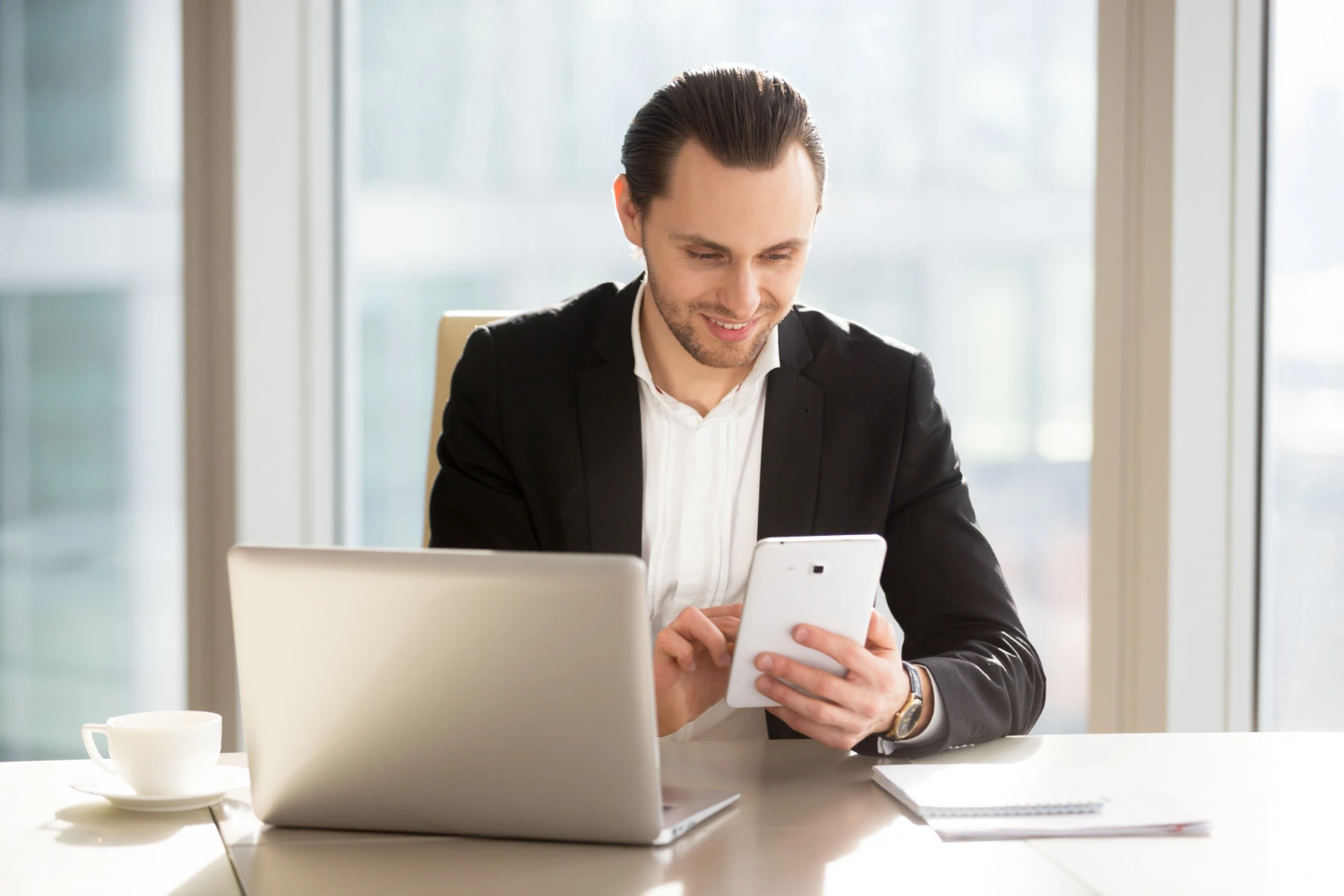 A person working on his tablet with a laptop, coffee, and some documents on top of his desk.