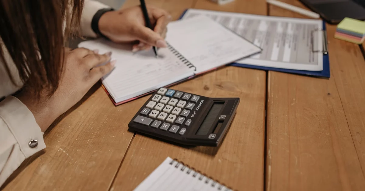 Woman preparing chart of accounts for small business using calculator and financial documents at desk.