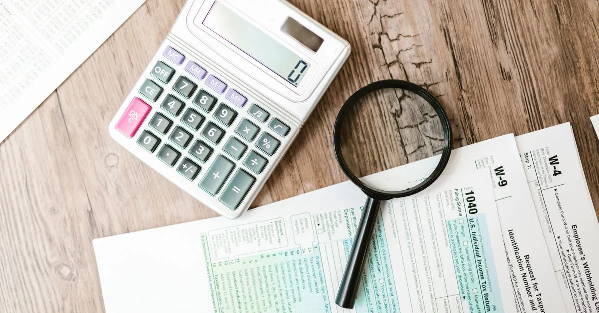 Calculator, magnifying glass, and tax forms on wooden desk for tax preparation cost assessment.