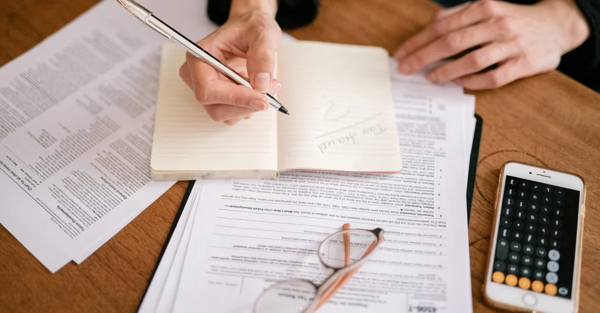 Person writing in notebook during corporate tax filing process with documents and calculator on desk.