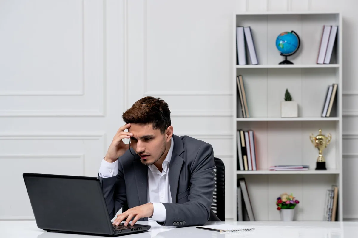 A man in a grey suit and headset rubs his forehead while frowning at his laptop in a white office.
